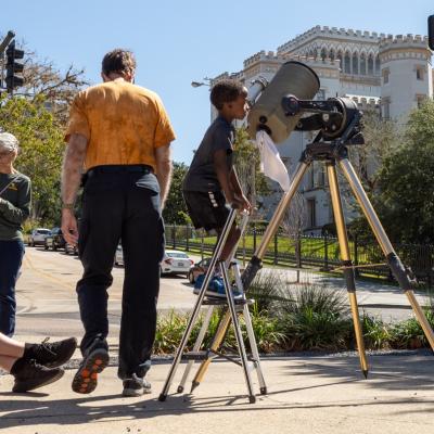A young boy uses a step stool to view the 2023 Annular Solar Eclipse from a solar scope standing on a tripod.