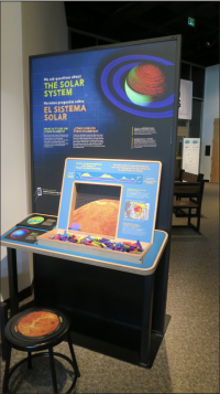 Single exhibition component, there is a large graphic that says The Solar System and a reading rail with a pixel puzzle that can be interacted with using small pieces of wood of various colors.