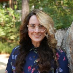 Peregrine Bratschi Headshot, woman standing among trees wearing a dark blue blouse with colorful butterflies