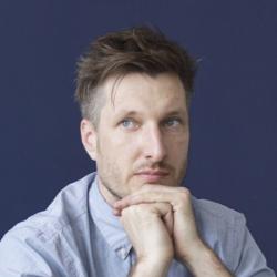 Headshot of Ethan Kruszka, individual with short hair looks into the distance while clasping his hands and resting his chin on them, shot from the shoulders up