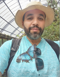 Paul Landa Headshot, man with beard smiles as a large blue morpho butterfly has landed on his shirt. He is wearing a hat, light blue shirt, and black backpack. He has his sunglasses clipped on his shirt collar.