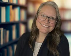 Individual from the shoulders up sitting in front of a blurred bookshelf. They are smiling and are wearing glasses and a black blazer. 