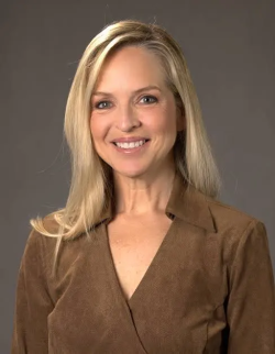 Closeup of an individual with shoulder-length blonde hair, smiling and wearing a brown top.