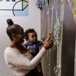 An adult holds an infant so they can feeling different textures on a wall that appear to be made from paper, tissue and other assorted crafting supplies.