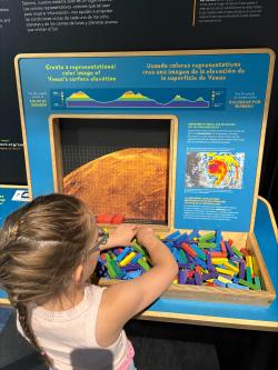 A young child sits at an exhibit component, there is a bin of small wood sticks of multiple colors and pixel puzzle where they are stacking these pieces.