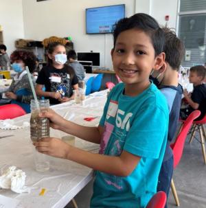 Child sitting at table in a classroom holding a jar with a straw in it doing hands-on science activity