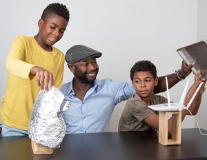 An adult and a child holds a metal baking sheet above the antennae of a Wi-Fi router on a block of wood, placed on a table. Another child holds a shell of tinfoil around a block of wood placed apart from the first block. 