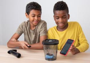 While sitting at a table, one child points a small transmitter at a vacuum-seal container that has a key finder inside, and another child holds a smartphone up to the other side of the container.