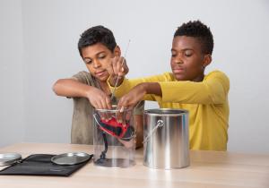 Two gallon-sized paint cans–one made out of clear plastic and the other metal– are side by side on a table. Behind them, two youths are seated at the table. Together they place a handheld, hand crank radio with a long antenna into the plastic paint can.