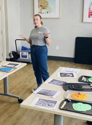 An educator stands at the front of a room with two long tables on either side. The tables have multiple trays with brightly colored clay rolled out. Next to each is an image of Moon craters.