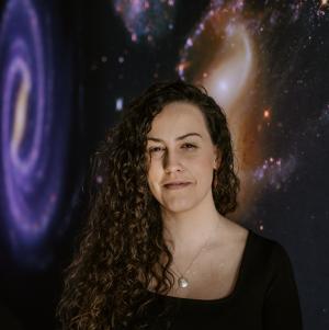Kaitlin Ehret Headshot - Women from shoulders up on a background with galaxy images, she has dark long curly hair over one shoulder and wears a dark shirt that blends into the background