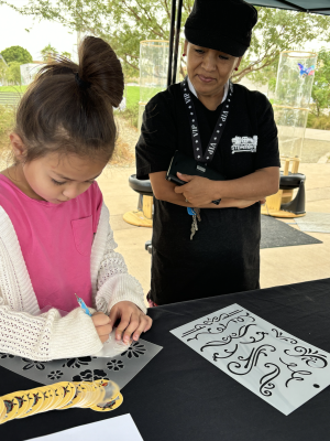 Child using a stylus and stencils to make tin art, behind the child is an individual wearing all black and watching.