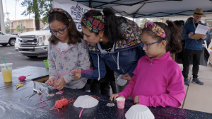 An adult stands between two young individuals at a table with various supplies for a hands-on activity.