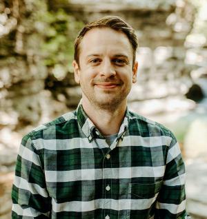 An individual stands in front of a blurred background, possibly outside. They are smiling, have short hair, and are wearing a checkered shirt.