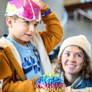 A standing child with a colorful paper hat on their head and an adult kneeling next to them. Both appear to be focused on a tabletop activity.