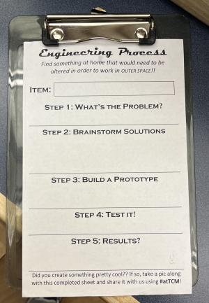 A small clipboard with white slip of paper that looks like a form not filled out, the top reads "Engineering Process" and 