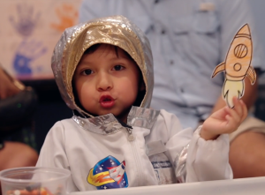 Child wearing a space suit costume at a community event holding a drawing of a rocket