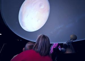 A child and caregiver gaze up at a planetarium screen showing a bright full Moon.