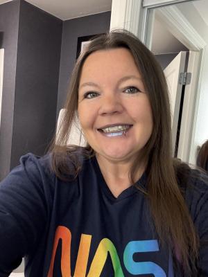 An individual stands in what appears to be a interior hallway, they are wearing a NASA shirt where the letters are rainbow colored. They are smiling and have shoulder-length hair.