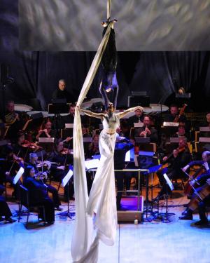 Two acrobats hang from a long line of fabric, one is in all black and the other in white. The two hang over a full orchestra playing on a stage.