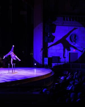 An acrobat poses in the shape of an X using only one hand on a small table or stool. A spotlight shines on them, creating a shadow on the wall that appears to be a person floating in space.