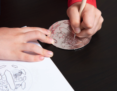 Hands Etch a Design into Foil as Part of the Moon Medallion Activity