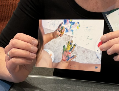 Hands holding up a What is Learning photo card showing toddler with paint