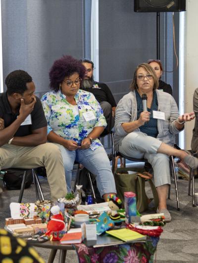 A diverse group sits and listens to one person, also seated, who has a mic. A small table is in front of the group and has many small items scattered on top.