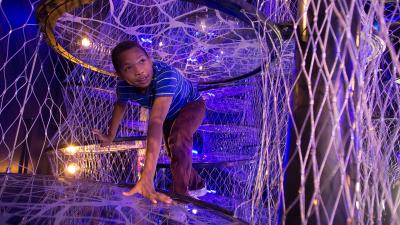 Young individual looks curiously as they crawl around an interconnected web-like climbing structure.