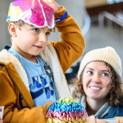 A standing child with a colorful paper hat on their head and an adult kneeling next to them. Both appear to be focused on a tabletop activity.