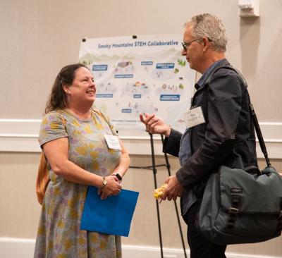 Two individuals have a conversation while standing in front of a poster on an easel. The poster reads "Smoky Mountain STEM Collaborative"