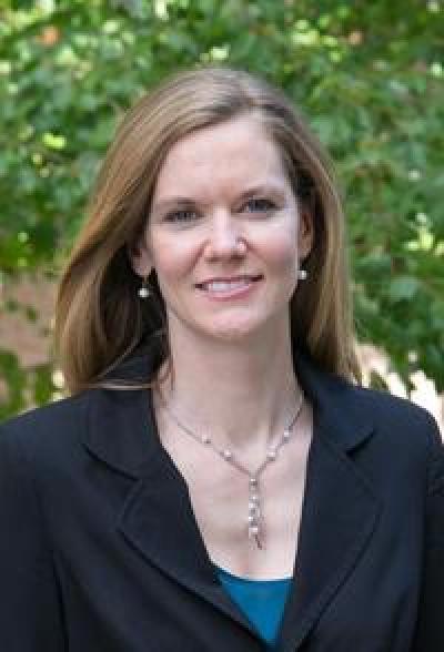 Headshot from shoulders up of individual with long brunette hair, standing outside in front of greenery and smiling