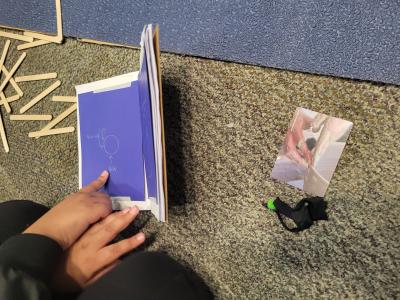 Hands point to the blue page of a small journal that has a What Is Learning card next to it.