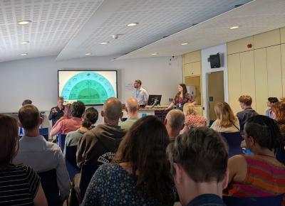 A large group watches a presentation, the current slide is a Decarbonization Infographic