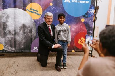 An adult kneels next to a child while, both are standing in front of a photo backdrop with a large Moon and other celestial objects and colors. 