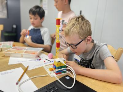 Three youths, two sit while one stands, at a table with trays of Lego bricks building robots.