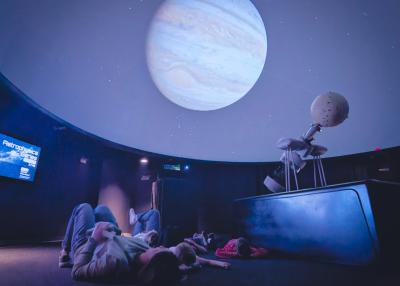 A child and caregiver gaze up at a planetarium screen showing Jupiter.