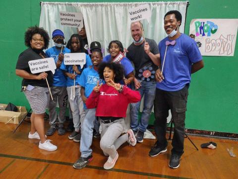 A group of kids and adults pose for the Communities for Immunity Photo Booth with handheld signs that say "Vaccines are Safe!"