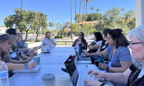 Group seated around a table outdoors, there are clear blue skies and palm trees. The group is working and conversing, focused on their task.