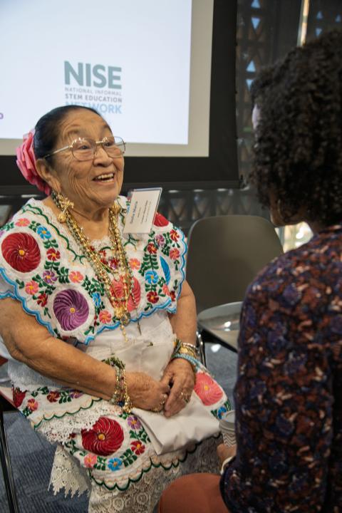 An elder sits in front of a projection screen while an individual leans into speaking to them.