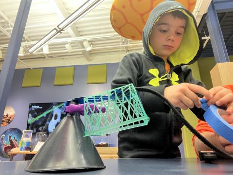A child stands at a table focused on creating a contraption with random items and tape.