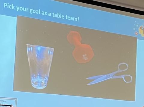 Projected slide reads "Pick your goal as a table!" with images of a clear drinking glass, small orange weight, and pair of scissors 