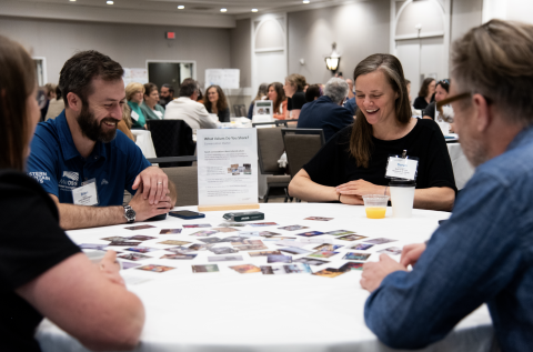 A group sits around a table, smiling, while reviewing many cards scattered across the top.
