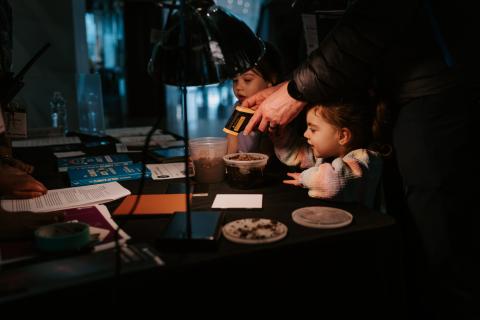 Two children stand at a table in a very dark space. There is a single light on the table, we can see some materials across the table and an adult hand holding a temperature gun aimed at some soil in a plastic container.