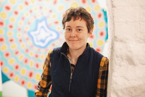 An adult wears a black vest over a flannel shirt, smiling and standing in front of some sort of spiral artwork.