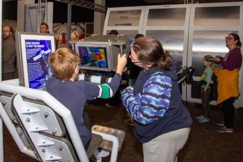 A youth sits in front of a monitor while and adult stands beside them. Both are focused on what appears to be a heat map on the screen, as the seated child engages with the screen's controls.