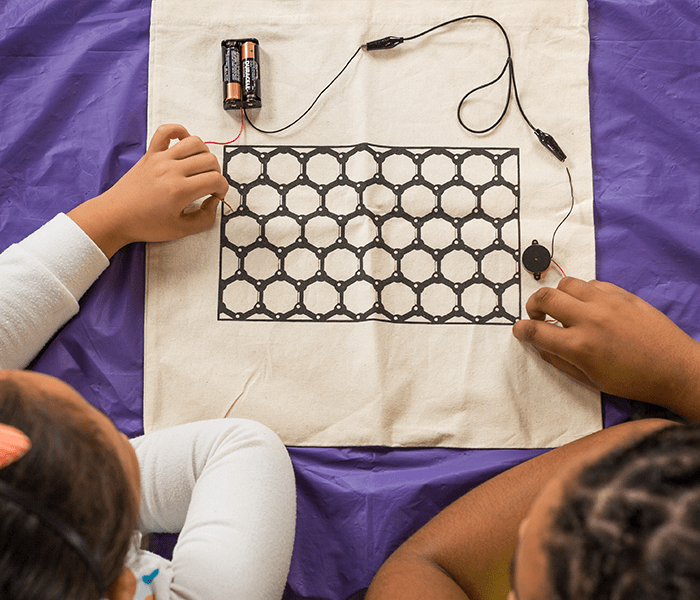 Children's hands exploring electrical conductivity with wires using cloth printed with graphene ink showing carbon structure model