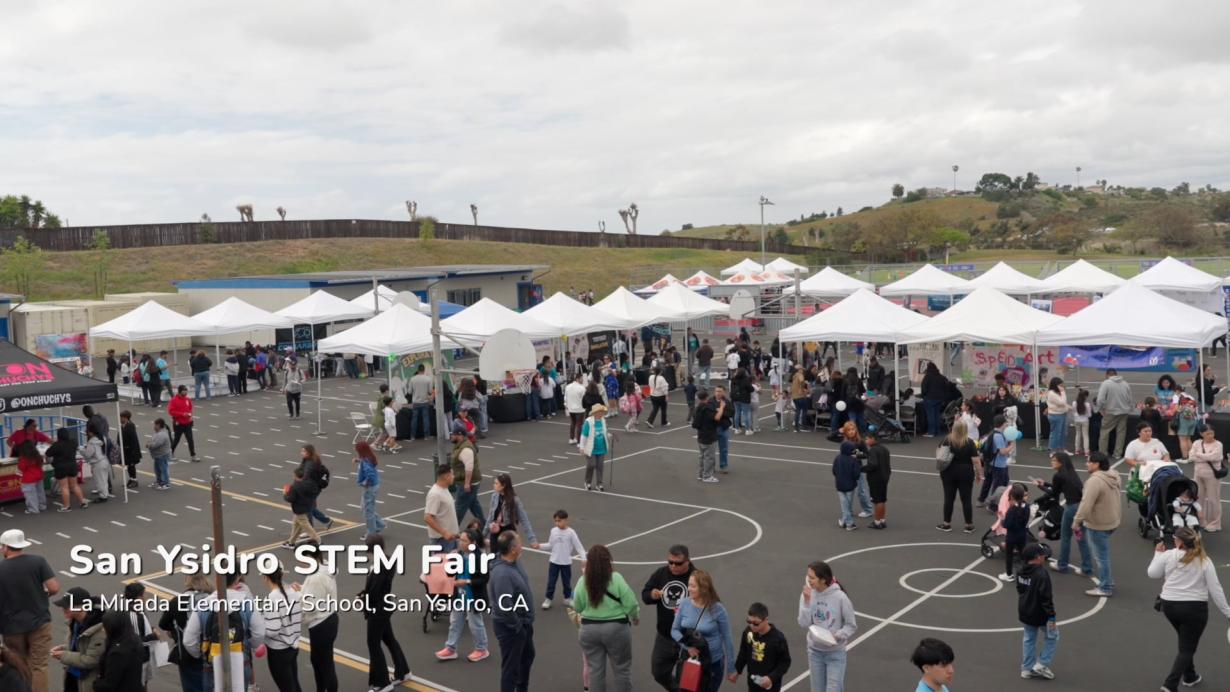Many event tents setup outside on a basketball court with a lot of people walking around the area.