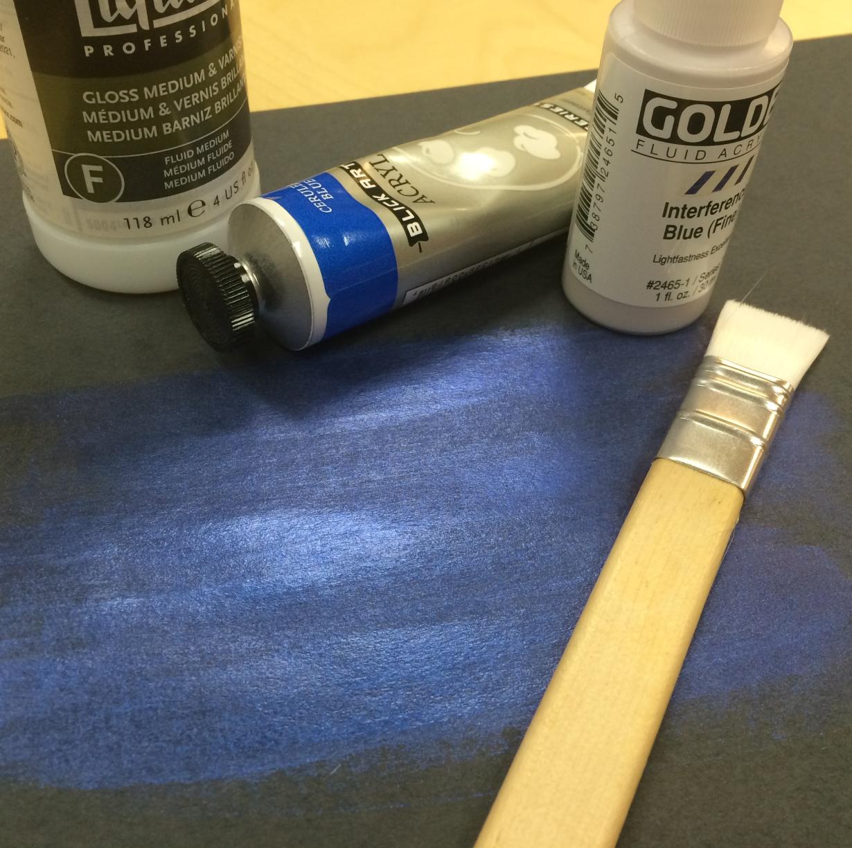 Paint tubes and a paint brush on top of paper colored with blue paint