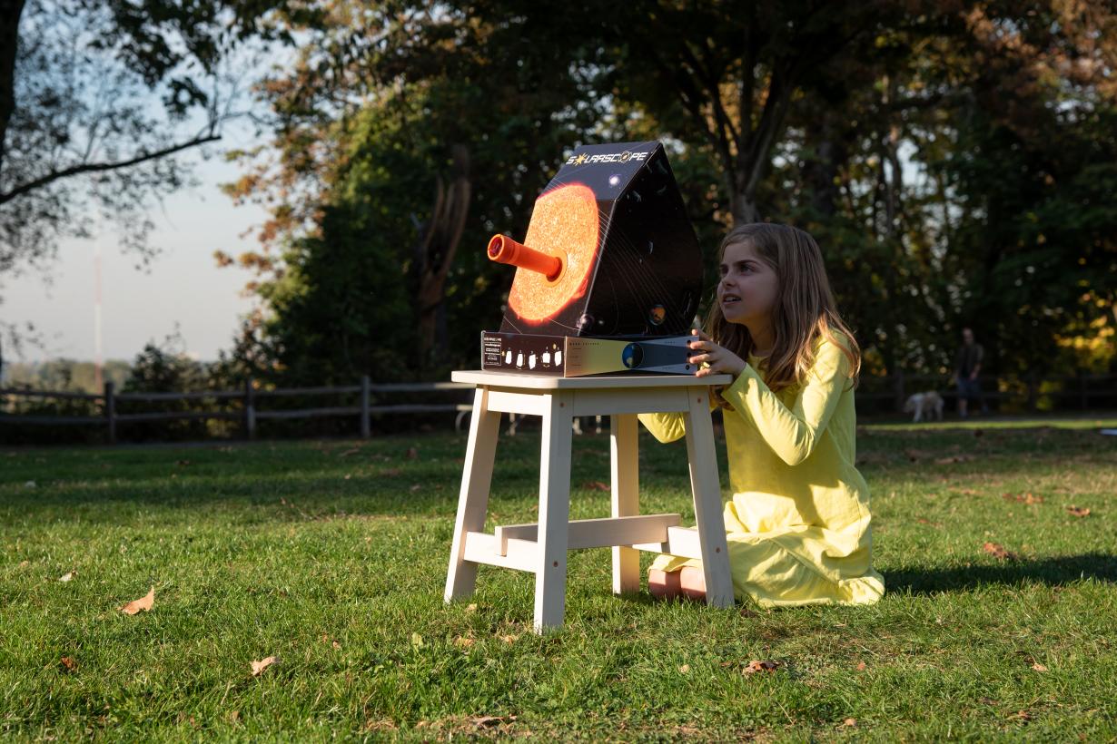 A young learner in a grassy field observes an image of the Sun being projected on her solarscope